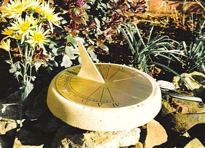 The round Jacobean brass sundial seated on a pale stone plinth in a rockery, its slim brass gnomon throwing a shadow across the engraved hour lines and numerals, with yellow chrysanthemums and grasses growing behind.