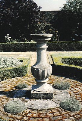 A Classic brass dial on a tall urn shaped Linford Manor pedestal at the centre of a brick paved circle, in a formal garden with clipped box beds around it.