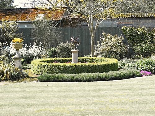 An Orbis armillary on a slim pedestal at the centre of a ring of clipped box hedging, on a long lawn with white planting and a bare tree behind it.