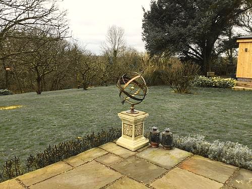 An Orbis armillary on a square stone plinth at the corner of a flagged terrace, looking out over a frosted lawn and orchard trees.