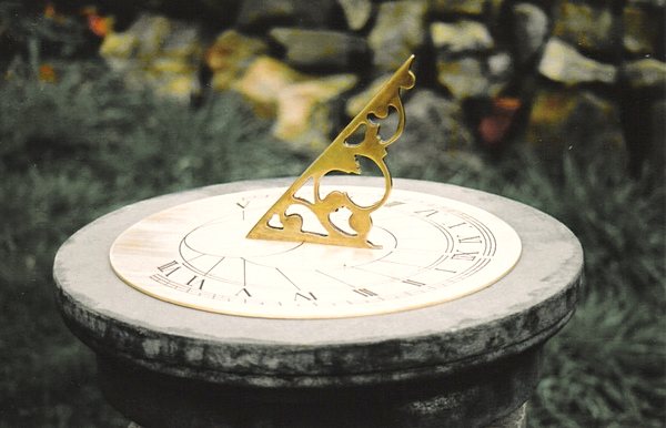 A pierced and scrolled brass gnomon standing on a round engraved dial plate on a stone pedestal, its shadow falling across the Roman numerals.