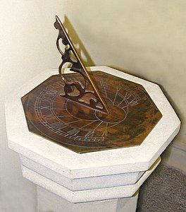 A Presentation dial made in an octagonal format, its weathered brass plate engraved with hour lines and numerals and fitted with a pierced scrolled gnomon, set on a stepped octagonal stone pedestal and photographed indoors against a plain wall.