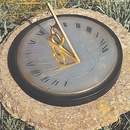 The Presentation sundial bedded into a rough stone block on a lawn, its darkened bronze-toned plate engraved with Roman numerals and hour lines inside a black rim, fitted with a decorative openwork brass gnomon that casts a shadow across the face.