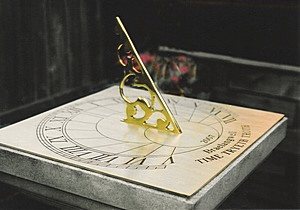A Presentation dial made in a square format, its pale engraved plate carrying Roman numerals, an inscription along the lower edge and a scrolled brass gnomon, photographed indoors on a dark surface with flowers behind.