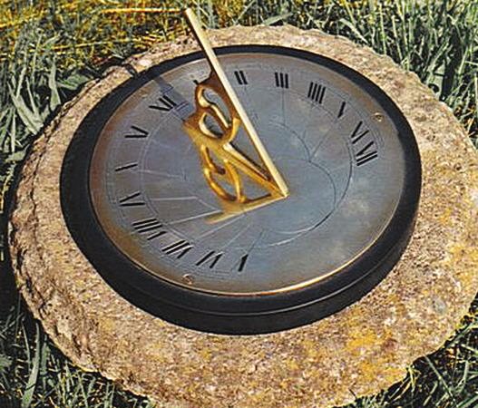 The Presentation sundial bedded into a rough stone block on a lawn, its darkened bronze-toned plate engraved with Roman numerals and hour lines inside a black rim, fitted with a decorative openwork brass gnomon that casts a shadow across the face.