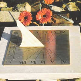 The square Regency brass sundial lying on a pale stone slab, its engraved Roman numerals running around three edges and fine hour lines fanning across the plate, with a plain brass gnomon casting a shadow and two orange gerbera flowers behind it.