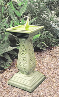 A Regency square sundial mounted on top of an ornate Venetian pedestal, both finished in green, standing on a gravel path with tall ferns and shrubs growing behind.