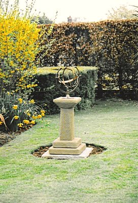 A Capricornus armillary on a small rustic stone pedestal set into a lawn, with forsythia in yellow flower and a beech hedge behind it.