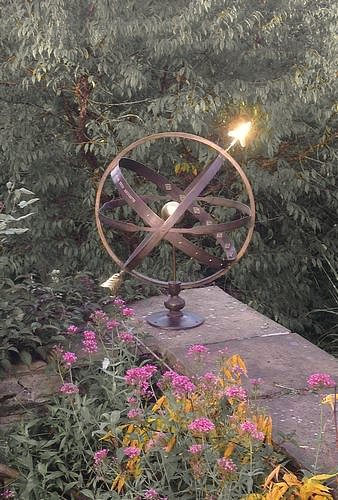 A bronze Solstice armillary on a dark pedestal against a brick garden wall, its arrow gnomon catching the low sun, with pink and yellow flowers in front of it.