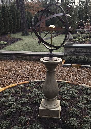A bronze Solstice armillary on a tall pale baluster pedestal standing in a gravel bed edged with low clipped box, with conifers behind it.