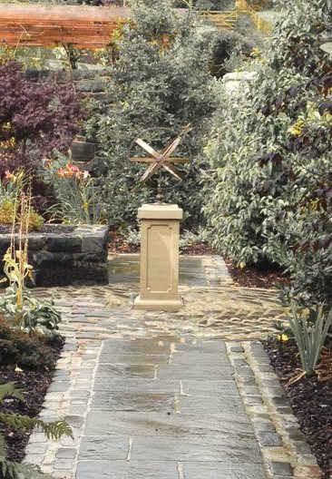 A bronze Solstice armillary on a plain square Tuscan plinth at the end of a stone flagged path running between planted borders.