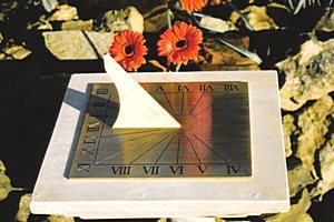 The square Regency brass sundial on its pale stone slab in front of a rockery, with two orange gerbera flowers behind, as shown on the catalogue page.