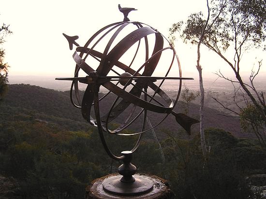 A large bronze Taurus armillary silhouetted against a hazy valley at sunset, with hillside trees behind it.