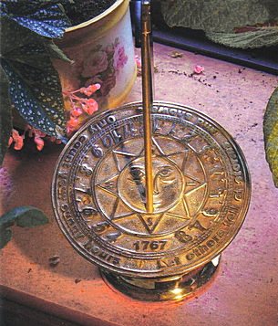 A close view of the Umbra's traditional Sunny Face dial plate in brass, a smiling sun in relief at the centre surrounded by a ring of engraved lettering and an engraved date, with the brass gnomon rod rising through the middle.
