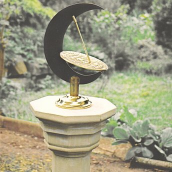 The Umbra upright sundial standing on an octagonal stone pedestal in a garden, a tall black crescent moon holding a small round engraved brass dial with a long brass gnomon rod passing through its centre, with lawn and shrubs behind.