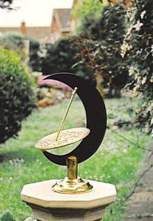 The Umbra upright sundial on its stone pedestal, the black crescent moon and brass dial seen against a garden lawn with trees and houses beyond.