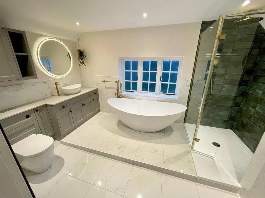Bathroom fitted by Telford Bathrooms with a freestanding bath under a window, brass floor standing tap, marble effect walls, a round backlit mirror and a dark green tiled shower