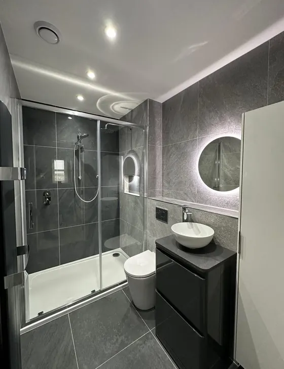 Shower with sliding glass doors, a round backlit mirror, black vanity and a vessel basin