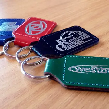 Four leather keyrings in navy, red, black and green laid out on a wooden desk, each blocked with a silver foil logo, including one reading Regent Garage.