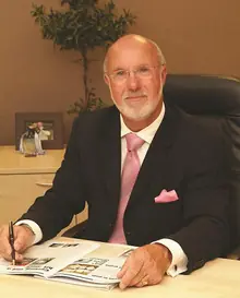 A portrait of a man with white hair and a white beard in a dark suit and pink tie, sitting at a desk with a pen in his hand and an open printed booklet in front of him.