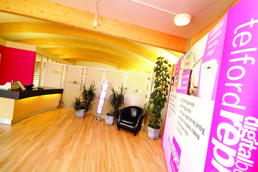 The Telford Repro reception: a wooden floor, a pale counter, potted plants and a tall magenta telford repro digital print banner against a yellow wall.