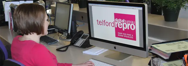 A designer at a desk in the Telford Repro studio working at a large iMac showing the telford repro digital bureau logo and the words short run print and design specialists, with a desk phone, keyboard and folders alongside.
