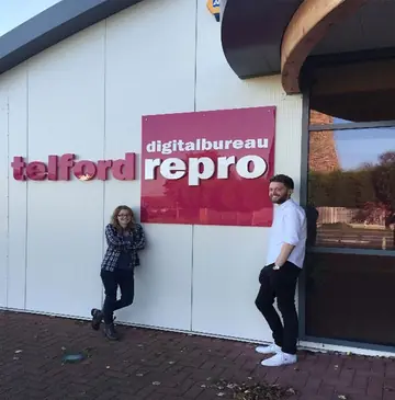 Two members of staff standing either side of the large telford repro digital bureau sign fixed to the outside wall of the building, one leaning against the wall in a checked shirt, one in a white shirt with hands in pockets.