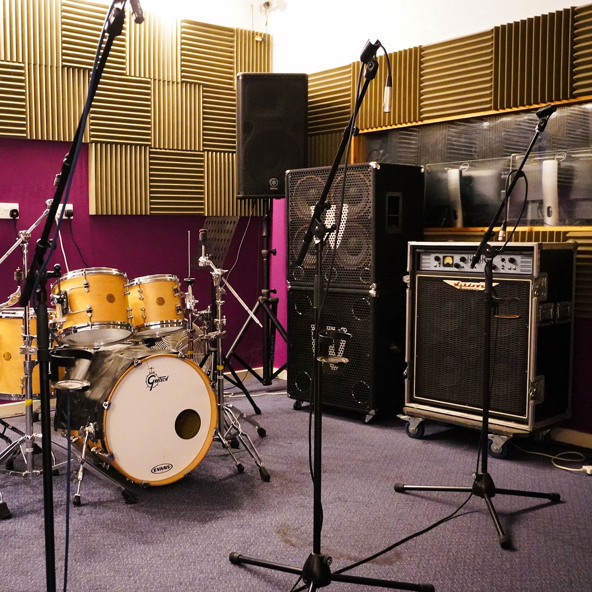 Rehearsal room with a natural maple Gretsch drum kit, bass rigs and gold acoustic foam against a deep magenta wall