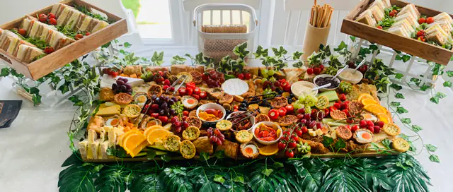 A long grazing table under a bright window, dressed with foliage and heaped with sharing food