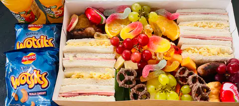 Layered sandwiches beside grapes, orange slices and pretzels on a grazing board
