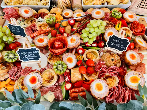 An overhead grazing spread with a small chalkboard reading Let us Graze, surrounded by eggs, salami and grapes