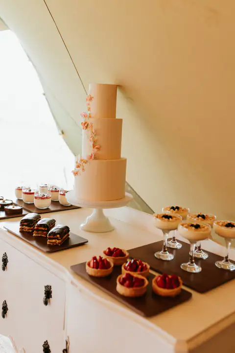 A blush wedding cake on a dessert table with tartlets, brownies and possets